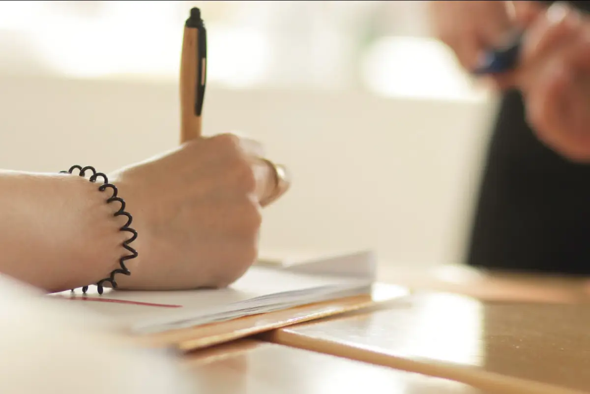 Nahaufnahme einer Person, die an einem Tisch mit einem Stift in ein Notizbuch schreibt. Auf dem Tisch liegen weitere Unterlagen und ein Stift. Im Hintergrund sitzt eine weitere Person und macht sich Notizen. Die ruhige Lernsituation vermittelt Aufmerksamkeit, Austausch und persönliches Lernen. Das Bild passt zum Thema Positive Feedbackkultur: Wertschätzung sichtbar machen und Entwicklung fördern und zeigt eine typische Situation aus einer Weiterbildung, in der Gedanken und Erkenntnisse festgehalten werden.