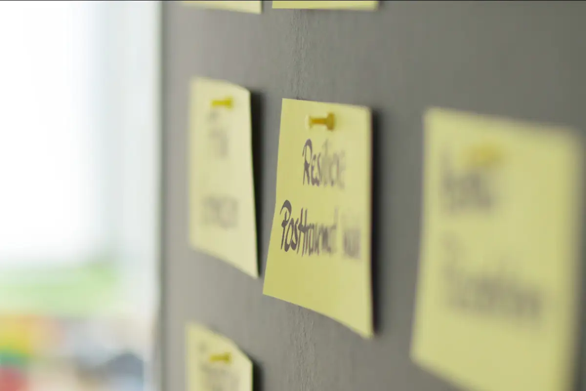 Nahaufnahme einer grauen Pinnwand mit mehreren gelben Haftnotizen. Die Notizen sind mit kurzen Stichworten beschriftet und mit kleinen gelben Pinnnadeln befestigt. Im Mittelpunkt steht eine Notiz mit dem Wort „Resilienz“. Weitere Notizen sind im Hintergrund zu sehen und teilweise unscharf. Die Pinnwand wirkt wie ein Arbeitsmaterial aus einem Seminar oder Workshop. Das Bild passt zum Thema Resilienz Workshop mit dem Resilienz Stärken Portfolio und zeigt eine Übung, bei der Gedanken und Erkenntnisse gesammelt und sichtbar gemacht werden